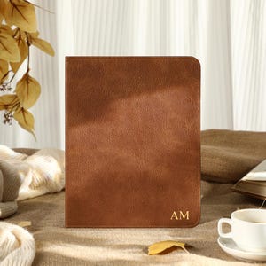May include: A brown leather journal, embossed with the gold initials "AM", is the central element. A white teacup and saucer are in the foreground, with a book and a beige sweater in the background. Dried leaves and sunlight contribute to the warm, inviting aesthetic.