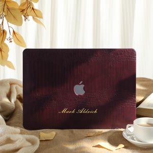 Può includere: Una custodia per laptop bordeaux con un motivo strutturato simile alla pelle di coccodrillo. Il logo Apple è visibile, insieme al nome "Mark Aldrich" in lettere dorate. Una tazza di caffè e un libro sono in primo piano.