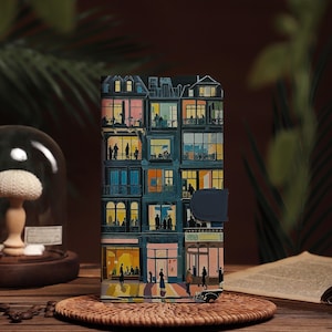 May include: A wallet featuring an artistic illustration of a multi-story building with lit windows and silhouettes of people. The color palette includes blues, oranges, and yellows, with a dark blue clasp. The wallet is displayed on a woven coaster.