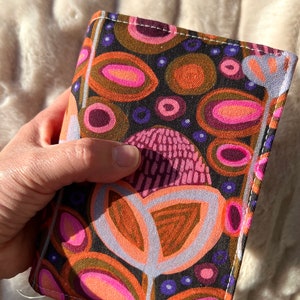 May include: A small, colourful fabric wallet with a geometric pattern of circles, ovals, and leaves in shades of brown, pink, orange, and purple. The wallet has a white border and is stitched with white thread.