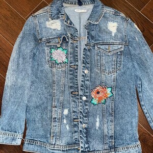 Può includere: Giacca di jeans blu chiaro con dettagli strappati e due toppe floreali. La giacca ha un colletto classico, chiusura con bottoni e due tasche anteriori. Le toppe floreali presentano sfumature di rosa, arancione e verde.