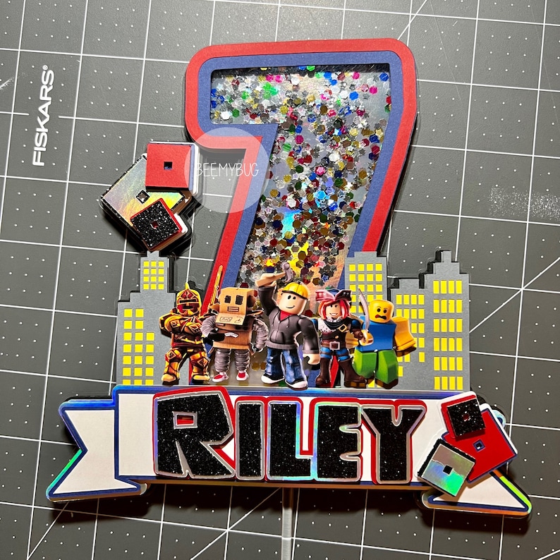May include: A 3D cake topper featuring the number 7 with a glittery background. The number is surrounded by a cityscape with Roblox characters. A banner at the bottom reads "RILEY 8".