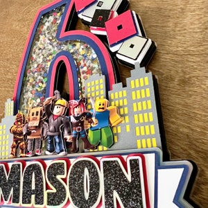 May include: A 3D cake topper featuring a large number six with glitter and confetti inside, a cityscape with yellow windows, and a row of Roblox characters. The name "MASON" is written in black glitter below the cityscape.