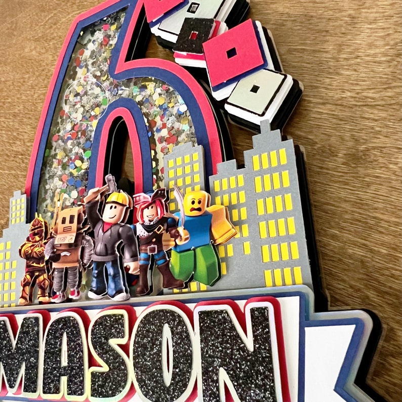 May include: A 3D cake topper featuring a large number six with glitter and confetti inside, a cityscape with yellow windows, and a row of Roblox characters. The name "MASON" is written in black glitter below the cityscape.