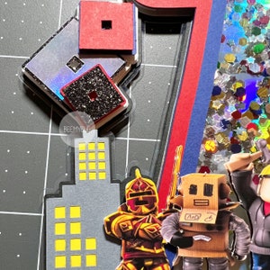 May include: A multi-layered birthday card featuring a large number seven, a skyscraper, and various Roblox characters. The characters include a knight, a robot, and a person wearing a construction helmet. The background is covered in colorful glitter.