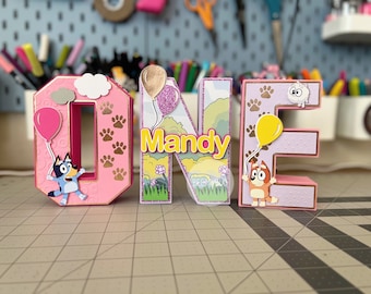 May include: Pink, purple and gold cardboard letters spelling "ONE" with a blue and white cartoon dog holding a pink balloon on the "O", a green and yellow cartoon dog holding a yellow balloon on the "E", and the name "Mandy" on the "N".
