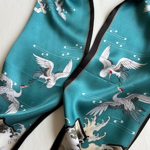 May include: A teal silk scarf featuring a repeating pattern of white cranes with red crowns in flight, accented by stylized white wave motifs and black border trim. The fabric has a smooth, lustrous finish with detailed illustrations of birds.