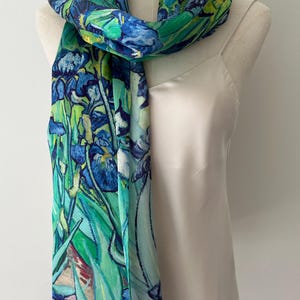 May include: A long rectangular scarf featuring a vibrant print of blue irises and green foliage. The fabric displays a painterly pattern with deep blue, light blue, and various shades of green. The scarf is draped around the neck of a mannequin.
