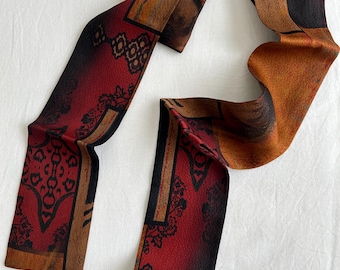 Pode incluir: Lenço estampado com tons de vermelho, castanho e preto, com desenhos geométricos. O lenço apresenta motivos detalhados e um acabamento texturizado. É exibido sobre uma superfície branca, realçando sua forma fluida e padrões intrincados.