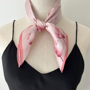 May include: A light pink square scarf with a floral tulip design in shades of pink and white. The scarf is tied around a mannequin's neck, showcasing its delicate fabric and pattern. The scarf is paired with a black top.