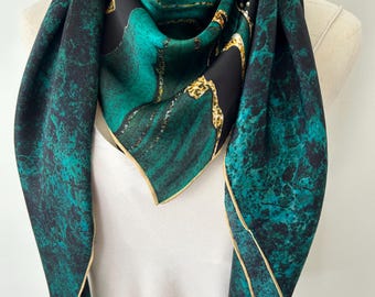 May include: A square scarf featuring a dark teal and black marbled pattern with gold-toned chain-link accents. The fabric has a smooth finish and is finished with a thin, light-colored border along the edges, draped around a mannequin neck.