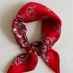 May include: A red bandana with a white paisley pattern, knotted in the center. The square scarf is made of a silky material and is folded to create a triangular shape. The bandana is a classic accessory.