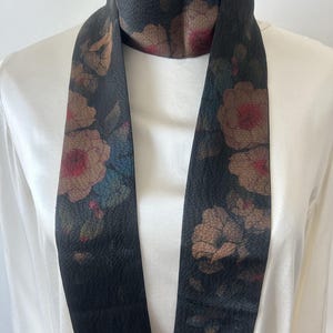 May include: A black silk scarf with a floral print in shades of brown, red, and blue. The scarf is long and narrow, and it is tied around a white mannequin's neck.