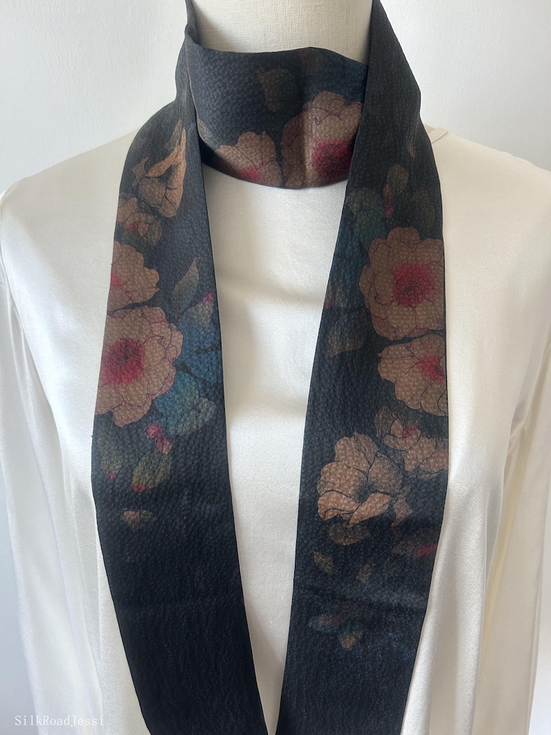 May include: A black silk scarf with a floral print in shades of brown, red, and blue. The scarf is long and narrow, and it is tied around a white mannequin's neck.