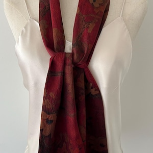 May include: A burgundy scarf with a floral pattern, draped over a white camisole. The scarf features a repeating design of brown and tan flowers and leaves. The scarf is knotted at the center, creating a stylish accessory.