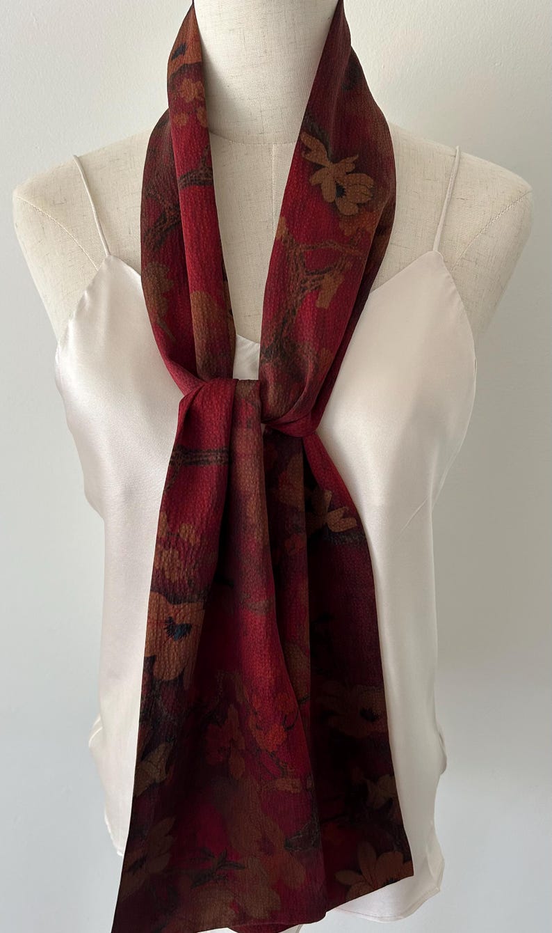 May include: A burgundy scarf with a floral pattern, draped over a white camisole. The scarf features a repeating design of brown and tan flowers and leaves. The scarf is knotted at the center, creating a stylish accessory.