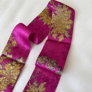 May include: A vibrant magenta silk scarf with a floral pattern in gold. The scarf features a repeating design of large flowers and cloud-like motifs. The fabric has a subtle sheen, suggesting a luxurious texture.