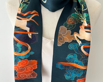 May include: A dark teal scarf featuring a repeating pattern of white deer with antlers, surrounded by stylized orange and blue clouds and flowing orange ribbons. The scarf is draped around a mannequin wearing a simple cream-colored top.