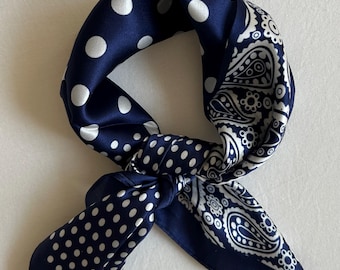 May include: A square scarf featuring a dark navy blue background with a white polka dot pattern on one side and a white paisley print on the other. The fabric has a smooth, satin-like finish and is tied into a loose knot, displaying both patterns.