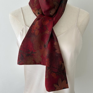 May include: A burgundy scarf with a floral pattern in shades of brown, orange, and green. The scarf is draped over a white mannequin, showcasing its design and texture. The scarf's fabric appears soft and lightweight.