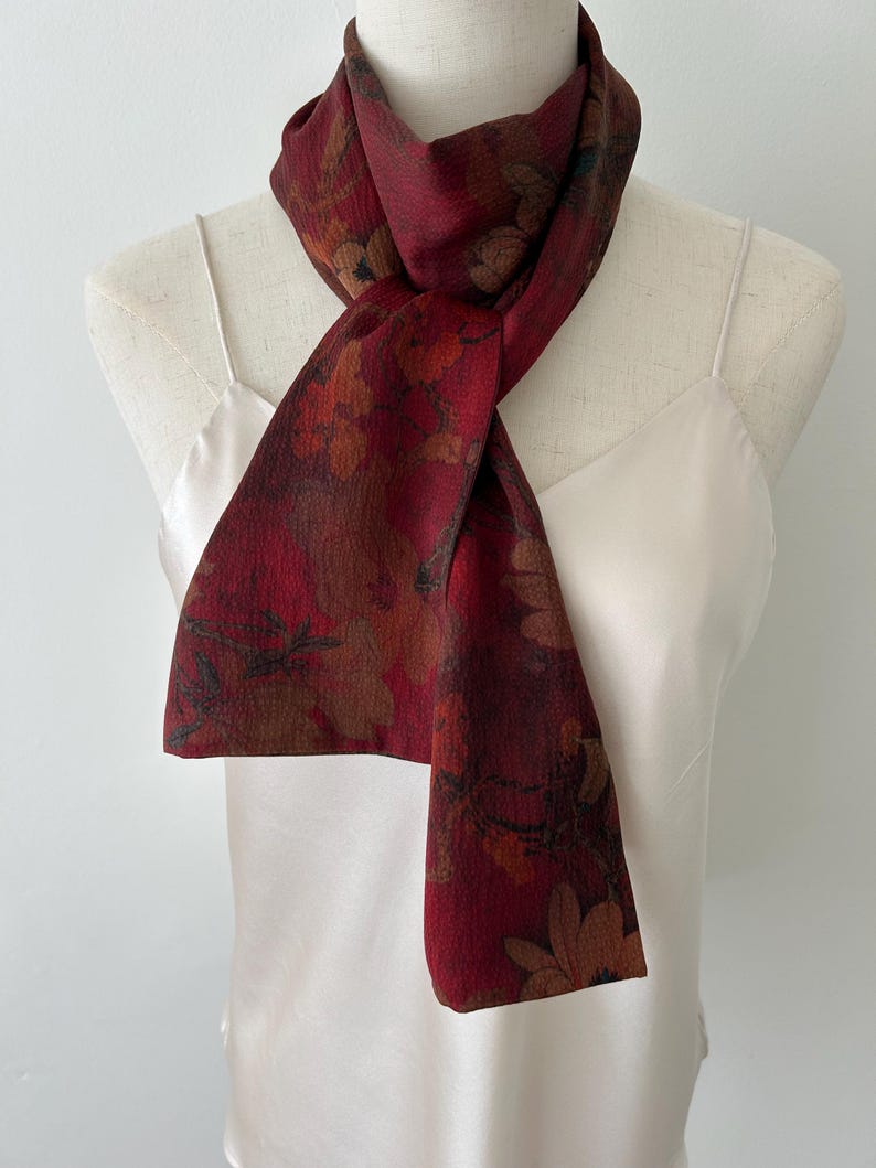 May include: A burgundy scarf with a floral pattern in shades of brown, orange, and green. The scarf is draped over a white mannequin, showcasing its design and texture. The scarf's fabric appears soft and lightweight.