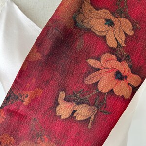 May include: A red floral scarf with a textured appearance. The scarf features large, stylized flowers in shades of orange and brown. The background is a deep red, creating a striking contrast with the floral design. The scarf is draped over a white garment.