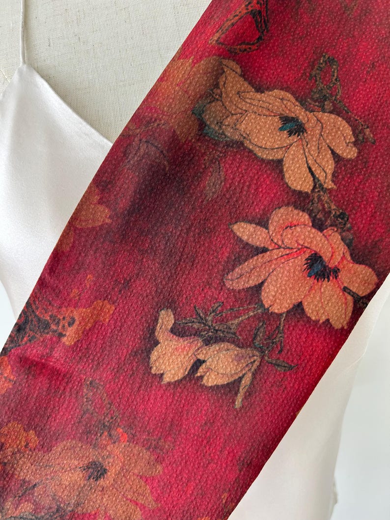 May include: A red floral scarf with a textured appearance. The scarf features large, stylized flowers in shades of orange and brown. The background is a deep red, creating a striking contrast with the floral design. The scarf is draped over a white garment.