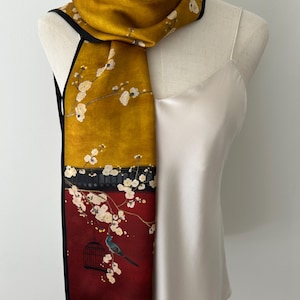 May include: A rectangular scarf featuring a mustard yellow upper section and a deep red lower section, separated by a black horizontal band. The design includes white cherry blossom branches, a small bird, and a black birdcage motif.