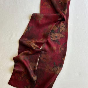 May include: A long, rectangular scarf in a deep red hue, adorned with a floral pattern in shades of brown and tan. The fabric appears to have a subtle texture, and the scarf is draped on a white surface.