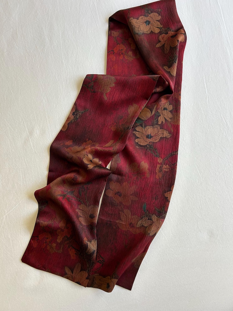 May include: A long, rectangular scarf in a deep red hue, adorned with a floral pattern in shades of brown and tan. The fabric appears to have a subtle texture, and the scarf is draped on a white surface.