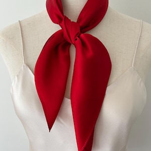 May include: A solid red square scarf tied in a simple knot around the neck of a mannequin. The scarf has a smooth, satin-like finish with a subtle sheen. The mannequin is wearing a plain, light-colored camisole with thin spaghetti straps.
