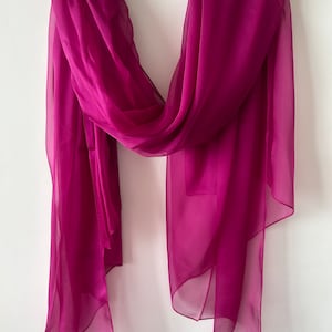 May include: A sheer magenta scarf draped over a wooden hanger. The scarf is made of a lightweight fabric and has a flowing, elegant drape.