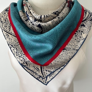 May include: A square silk scarf featuring a detailed paisley pattern in navy blue and cream, accented by a solid teal border and a vibrant red stripe. The scarf is folded and draped to display the intricate design and contrasting color palette.