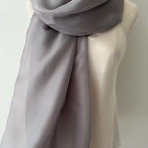 May include: A solid grey scarf with a smooth, semi-sheer texture draped over a cream-colored sleeveless top. The scarf features a soft, flowing drape and a consistent matte finish, providing a neutral accessory that complements the light-colored garment.