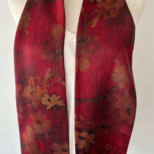May include: A red scarf with a floral pattern. The scarf has a textured surface and is draped over a white top. The floral design features brown and tan flowers and branches against a deep red background.