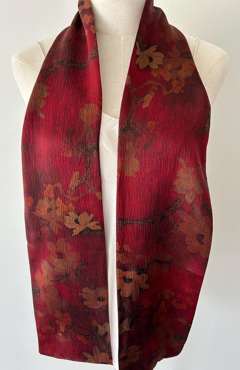 May include: A red scarf with a floral pattern. The scarf has a textured surface and is draped over a white top. The floral design features brown and tan flowers and branches against a deep red background.