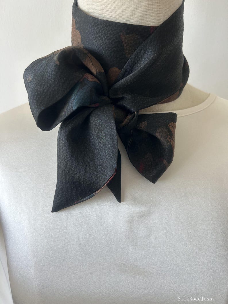 May include: A black silk scarf with a floral print. The scarf is tied around a mannequin's neck in a bow.