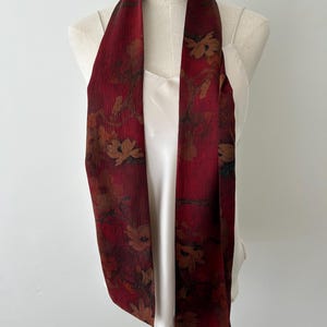 May include: A long, burgundy scarf with a floral pattern in shades of brown and tan. The scarf is draped over a white top on a mannequin. The scarf's fabric appears to be lightweight and flowing, suitable for adding a touch of elegance to an outfit.