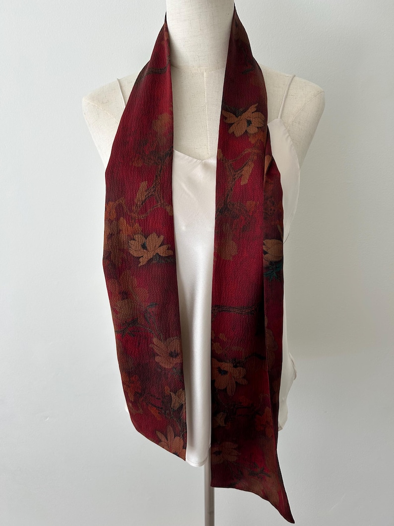 May include: A long, burgundy scarf with a floral pattern in shades of brown and tan. The scarf is draped over a white top on a mannequin. The scarf's fabric appears to be lightweight and flowing, suitable for adding a touch of elegance to an outfit.