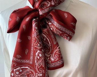 May include: A red paisley patterned silk scarf tied in a bow around a white collared shirt.