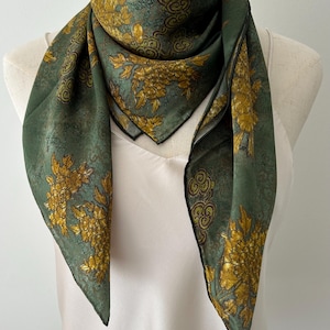May include: A square scarf in a muted sage green hue featuring a prominent gold floral pattern and dark swirling motifs. The fabric has a smooth texture with a thin black border along the edges, draped around the neck of a mannequin.