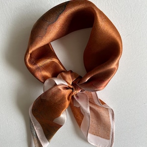 May include: A square silk scarf in shades of copper, brown, and cream, knotted in the center. The scarf has a subtle sheen and a delicate floral pattern along the edges. The scarf is a versatile accessory.