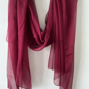 May include: A burgundy sheer scarf hanging on a wooden hanger. The scarf is made of a lightweight, flowing fabric.