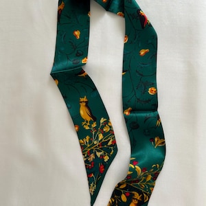 May include: A dark teal satin scarf with a repeating pattern of yellow birds, red berries, and yellow flowers. The scarf is long and narrow, with pointed ends. The design is set against a backdrop of dark green vines and leaves.