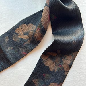 May include: A black silk scarf with a floral print in shades of brown, pink, and green. The scarf is tied in a knot and the ends are hanging down.