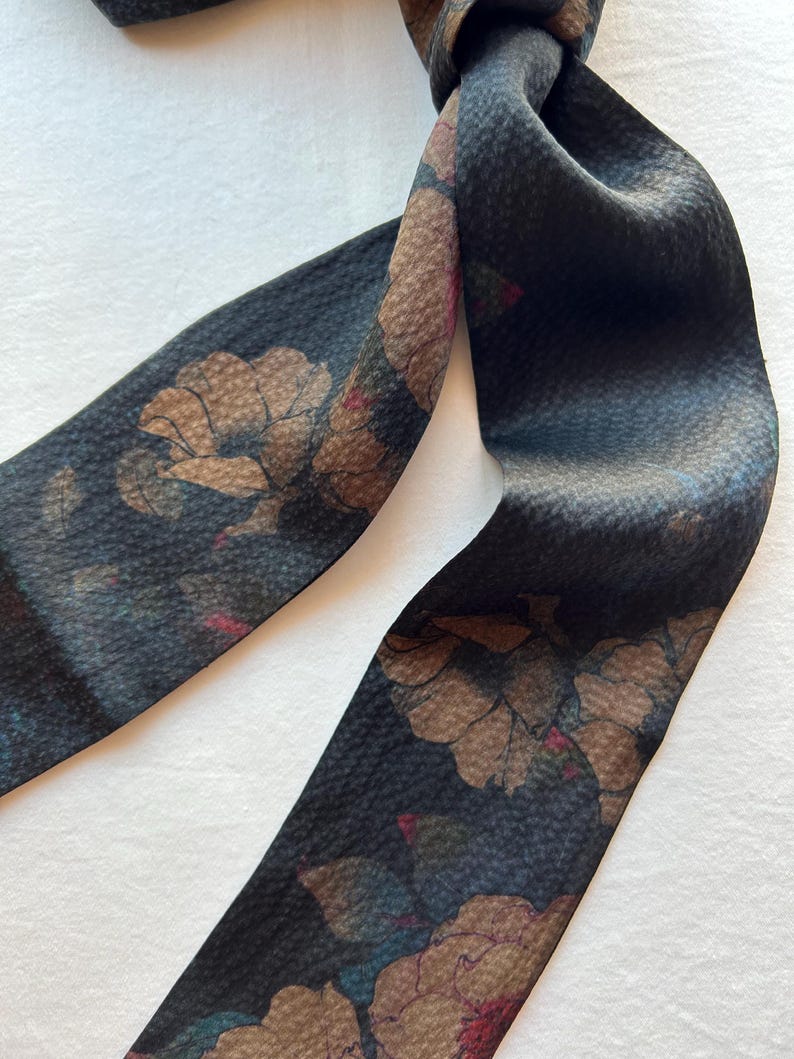 May include: A black silk scarf with a floral print in shades of brown, pink, and green. The scarf is tied in a knot and the ends are hanging down.