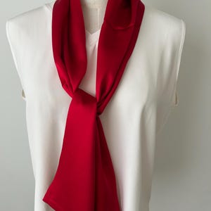 May include: A vibrant red scarf is draped around a white top. The scarf is knotted at the neck and hangs down, showcasing its smooth texture and rich color. The scarf adds a pop of color to the neutral background.