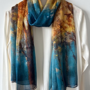 May include: A sheer scarf with a nature-inspired design. The scarf features a blend of colors, including teal, yellow, and brown, creating a watercolor effect. The scarf is draped over a white shirt on a wooden hanger.