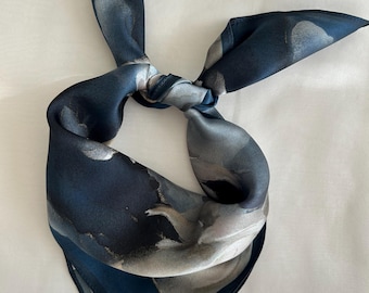 May include: A square silk scarf with a watercolor-like design in shades of navy blue, gray, and white. The scarf is knotted at the top, showcasing the flowing fabric and color variations. The edges are neatly finished.