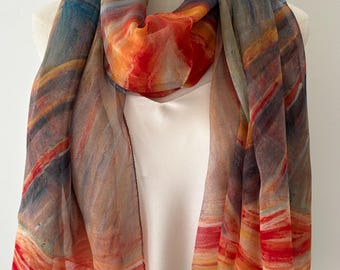 May include: A sheer scarf with a vibrant abstract design. The scarf features a blend of colors, including orange, red, blue, and brown, creating a watercolor effect. The scarf is draped over a white mannequin, showcasing its flowing fabric.